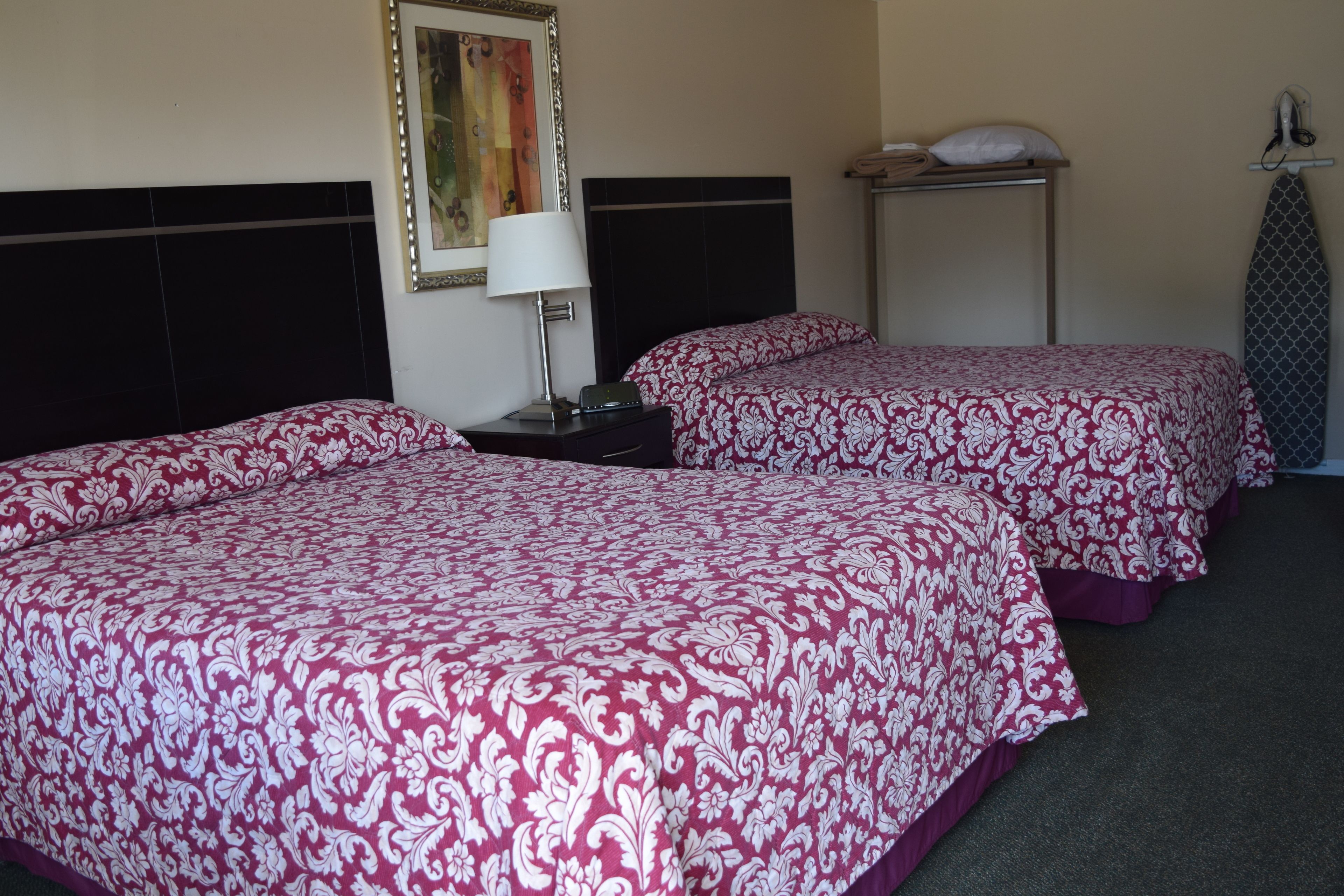 A hotel room with two beds, a nightstand, a lamp, a framed picture, a shelf with a pillow, and an ironing board.
