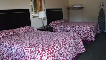 A hotel room with two beds, a nightstand, a lamp, a framed picture, a shelf with a pillow, and an ironing board.