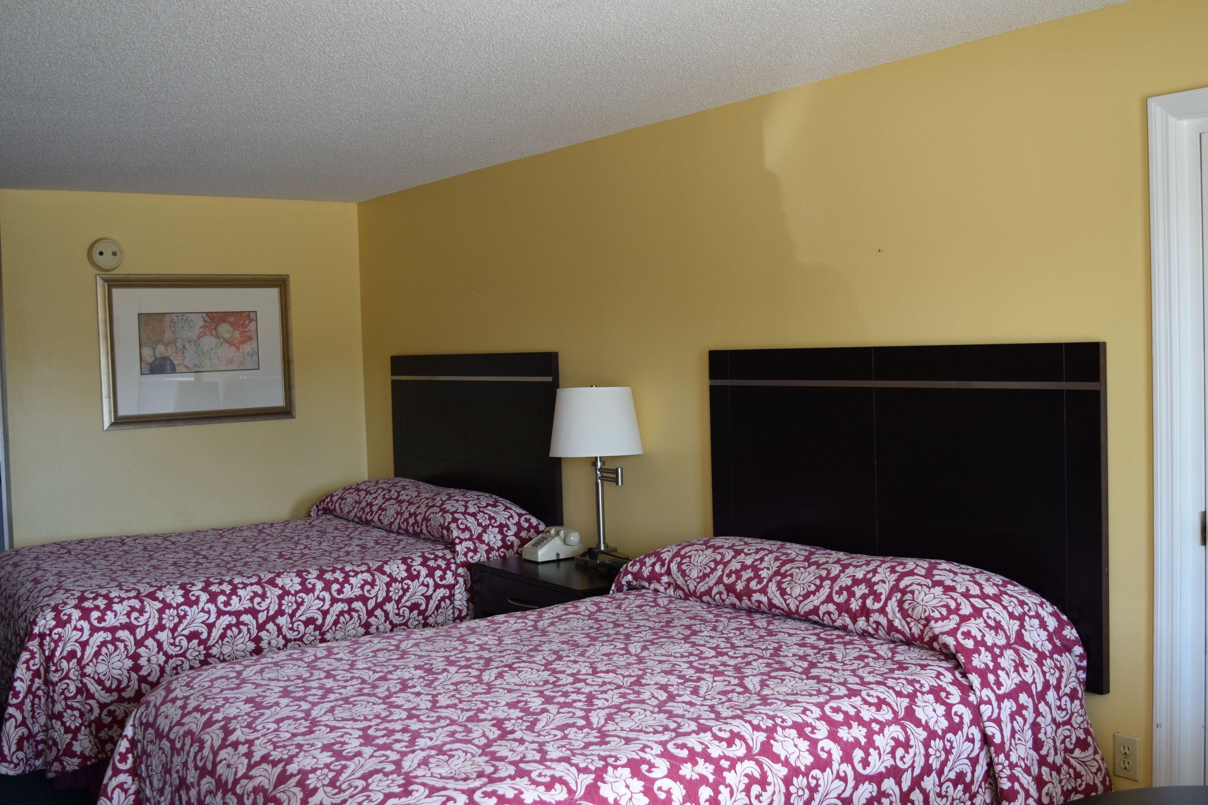A hotel room with two beds, each with a patterned bedspread, a bedside table with a lamp, and a framed picture on the wall.