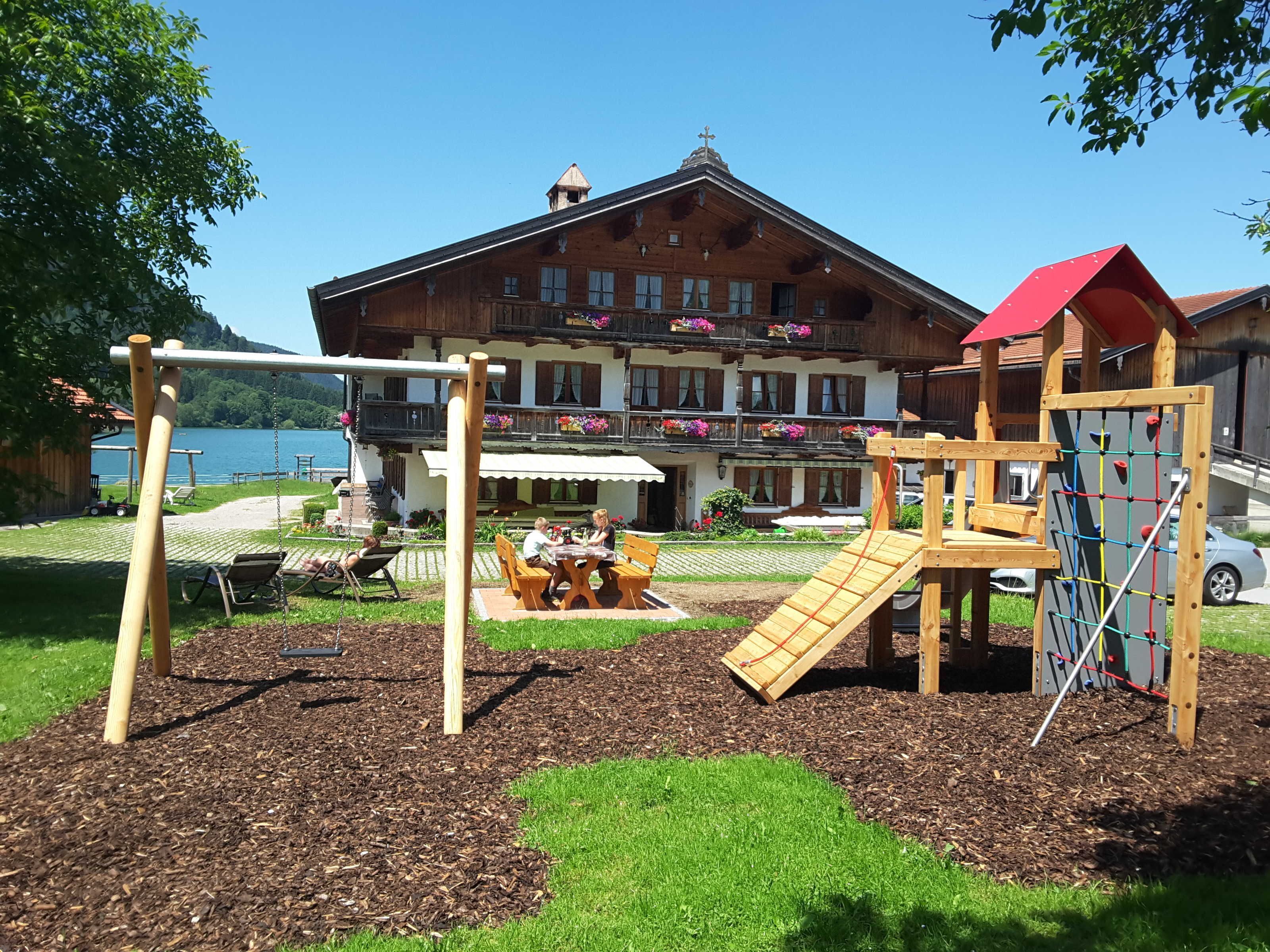 Un terrain de jeux avec des balançoires et des structures d’escalade, une table avec des chaises, et un bâtiment avec des balcons et des jardinières en arrière-plan.
