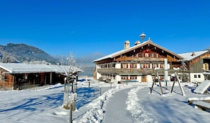 A traditional wooden building with a balcony, a snow-covered path, and a mountain in the background. - Vacation apartment Alpenrose - The Anderlbauer by the lake (Schliersee)