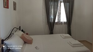 A neatly made bed with pillows and a remote control, a chandelier, and curtains on a window. - Il Silenzio (first floor) Magarikari South Crete (Magarikari)