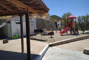 A playground with a red slide and swings, a barbecue area, and a stone wall. - Il Silenzio (first floor) Magarikari South Crete (Magarikari)