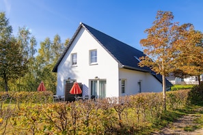 Ein weißes Gebäude mit dunklem Dach, zwei Fenstern und einer Tür. Vor der Fassade stehen ein roter Sonnenschirm und ein Tisch mit Stühlen.