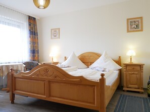 Una cama de madera con ropa de cama blanca, dos mesitas de noche con lámparas, una silla y una ventana con cortinas.