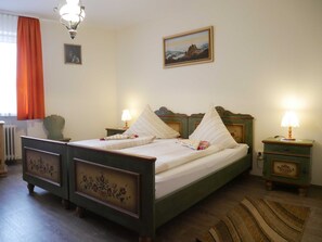 A neatly arranged bedroom with a bed, two bedside tables, a nightstand, a lamp, a framed picture on the wall, and a window with curtains.