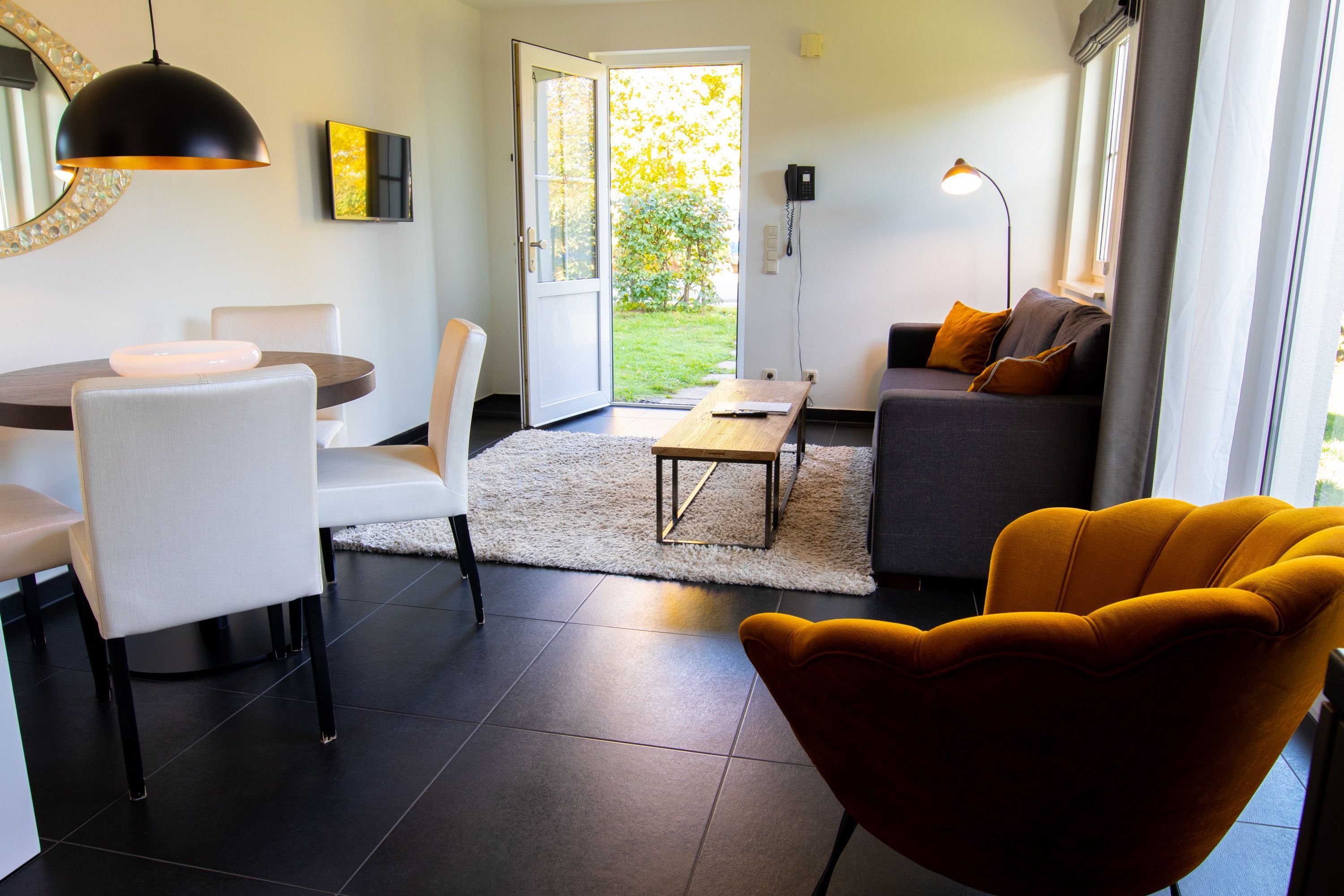 Una sala de estar moderna con comedor, un sofá con cojines, una silla amarilla y vistas a un jardín a través de un ventanal.