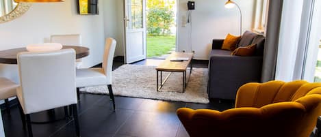 Una sala de estar moderna con comedor, un sofá con cojines, una silla amarilla y vistas a un jardín a través de un ventanal.