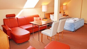 Habitación de hotel con mesa de vidrio, sofá de cuero rojo, sillas blancas, cama con colcha azul y tragaluz.