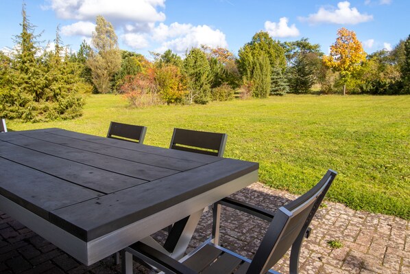 Una mesa de comedor con sillas, sobre una zona empavedada con un campo de césped y árboles al fondo.