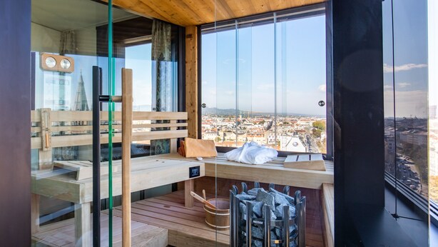 Una camera moderna con una panca in legno, un tavolo in legno con asciugamani, vista sulla città e area sauna con un catino e pietre.