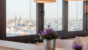 Una stanza con vista sulla città, un divano bianco, un secchio di metallo con fiori e una bottiglia su un tavolo.