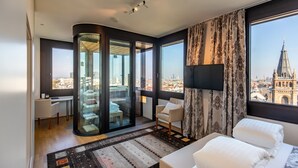 Una camera d'albergo moderna con una grande finestra che offre vista sulla città, un pannello di vetro che separa la zona living dal balcone, e un’accogliente area salotto con divano e poltrone.