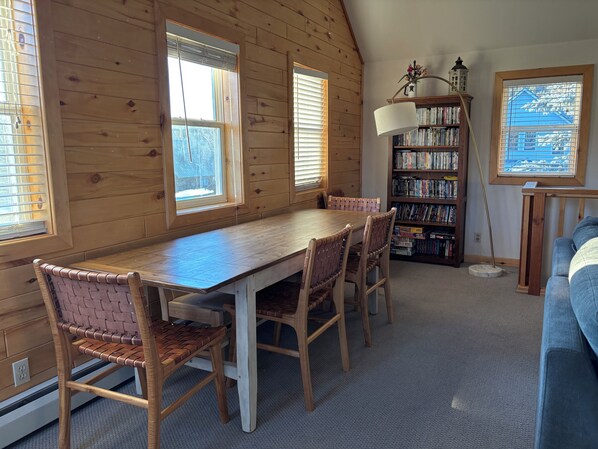 A wooden-paneled room with a dining table, chairs, a bookshelf, and a window with blinds.