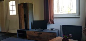 A room with a wooden cabinet, a television on a stand, a small table with a vase, and a window with curtains. - House 2 - Holiday home family Acker (Lychen)
