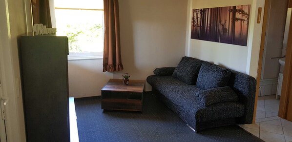 A small room with a sofa, a small table with a vase, a window with curtains, and a wall with a forest painting.