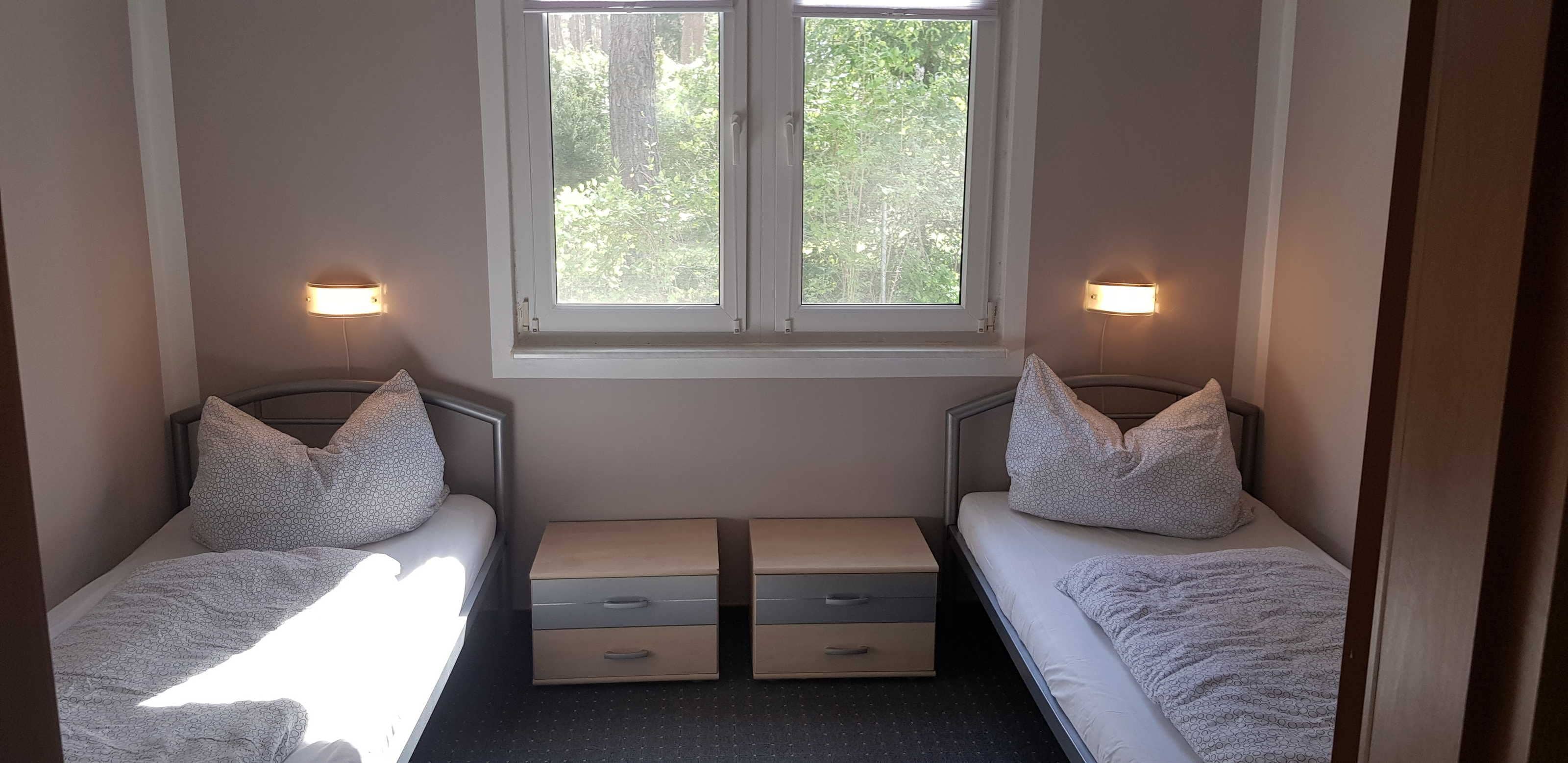 A small, well-lit bedroom with two single beds, each with a pillow and a bedside table with drawers.