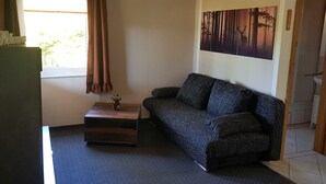 A small, furnished room with a sofa, a small wooden table with a vase, a window with curtains, and a wall-mounted painting. - House 2 - Holiday home family Acker (Lychen)
