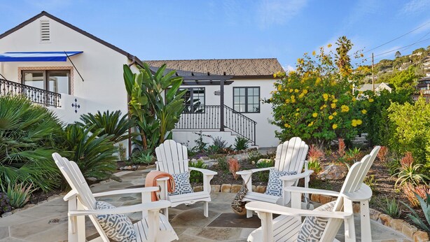 Un patio con sedie Adirondack bianche e pavimento in pietra, circondato da un giardino con diverse piante e una casa sullo sfondo.