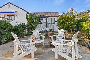 Une terrasse dotée de chaises Adirondack blanches et d’un sol en pierre, entourée d’un jardin où poussent différentes plantes, avec une maison en arrière-plan.