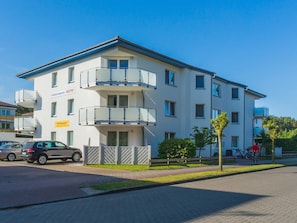 Un immeuble résidentiel à plusieurs étages, doté de balcons, d’un espace de stationnement et d’un petit arbre devant l’entrée.