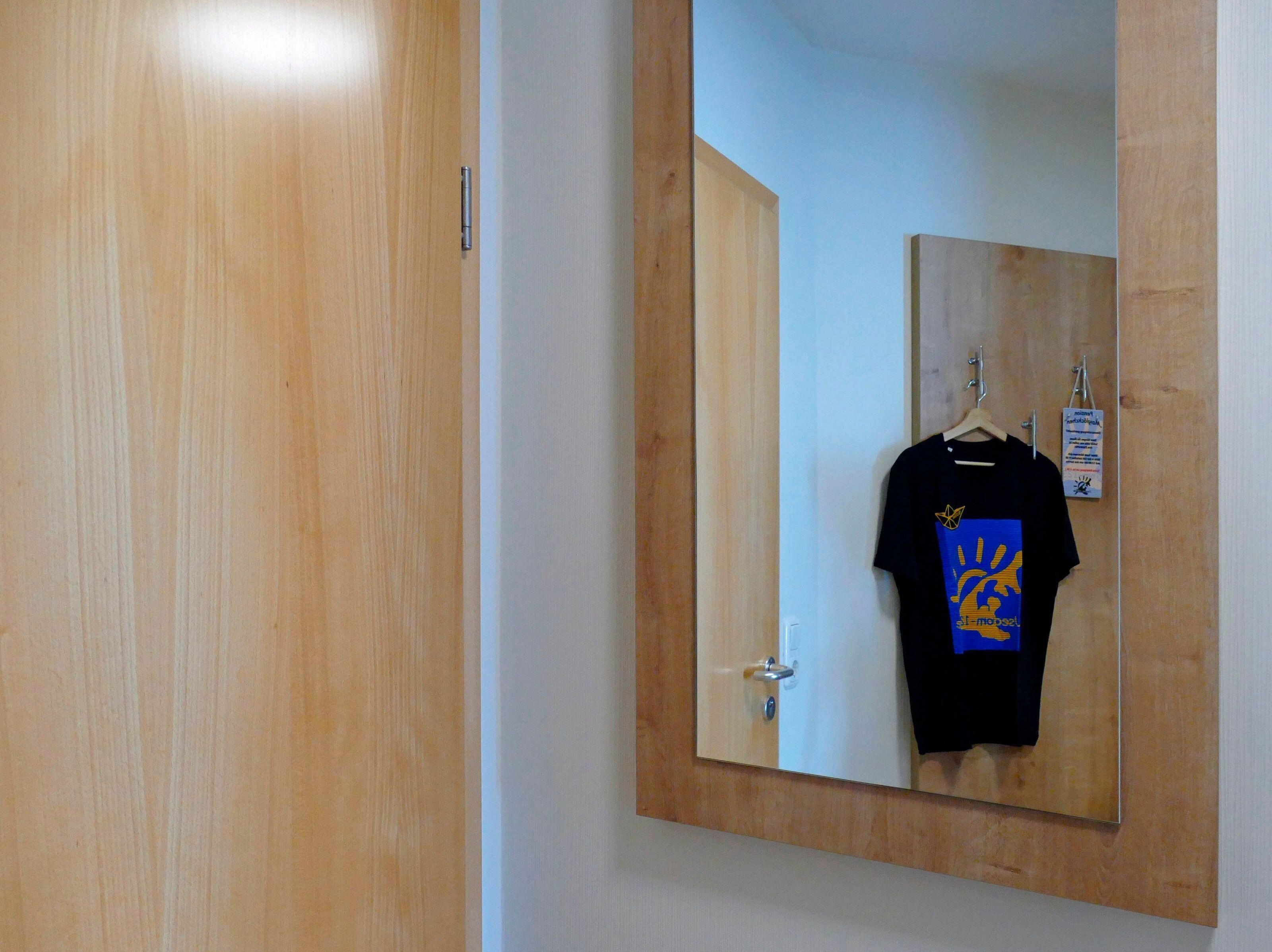 A wooden door with a mirror reflecting a black t-shirt hanging on a wall-mounted hanger.