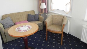 Huoneessa on sohva, tuoli, pieni pöytä, jonka päällä on kukkakuviollinen pöytäliina, lamppu, ikkuna, jossa on verhot, ja patteri.