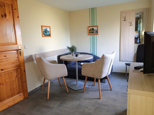 Een knus, klein kamertje met een houten deur, een ronde tafel met twee stoelen en een kleine plant in een pot. Er hangen twee ingelijste foto's aan de muur en een spiegel die een deel van de kamer weerspiegelt.