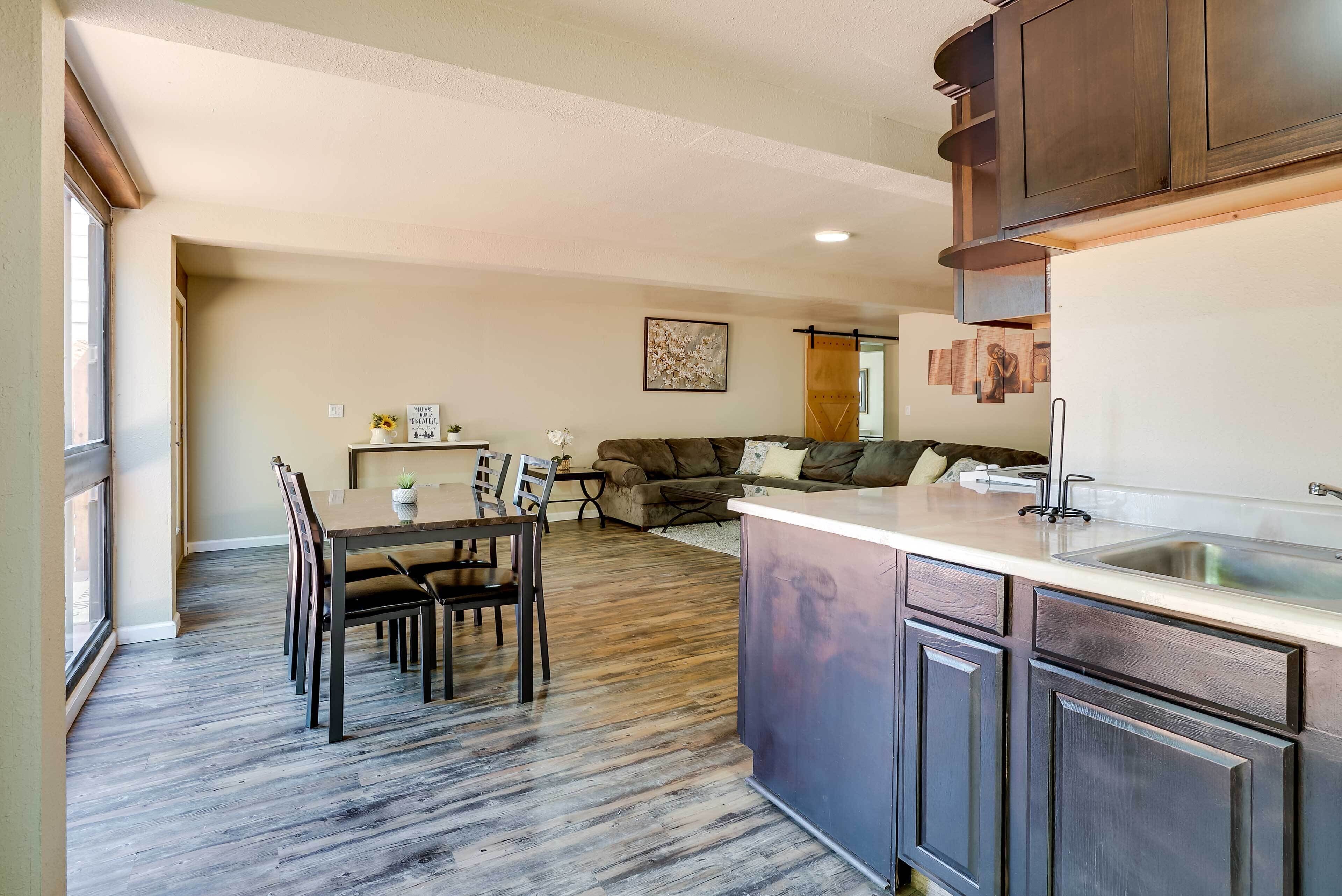 A modern kitchen with dark wood cabinets, a dining area with a wooden table and chairs, and a living area with a sofa and cushions.