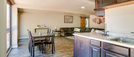 A modern kitchen with dark wood cabinets, a dining area with a wooden table and chairs, and a living area with a sofa and cushions.