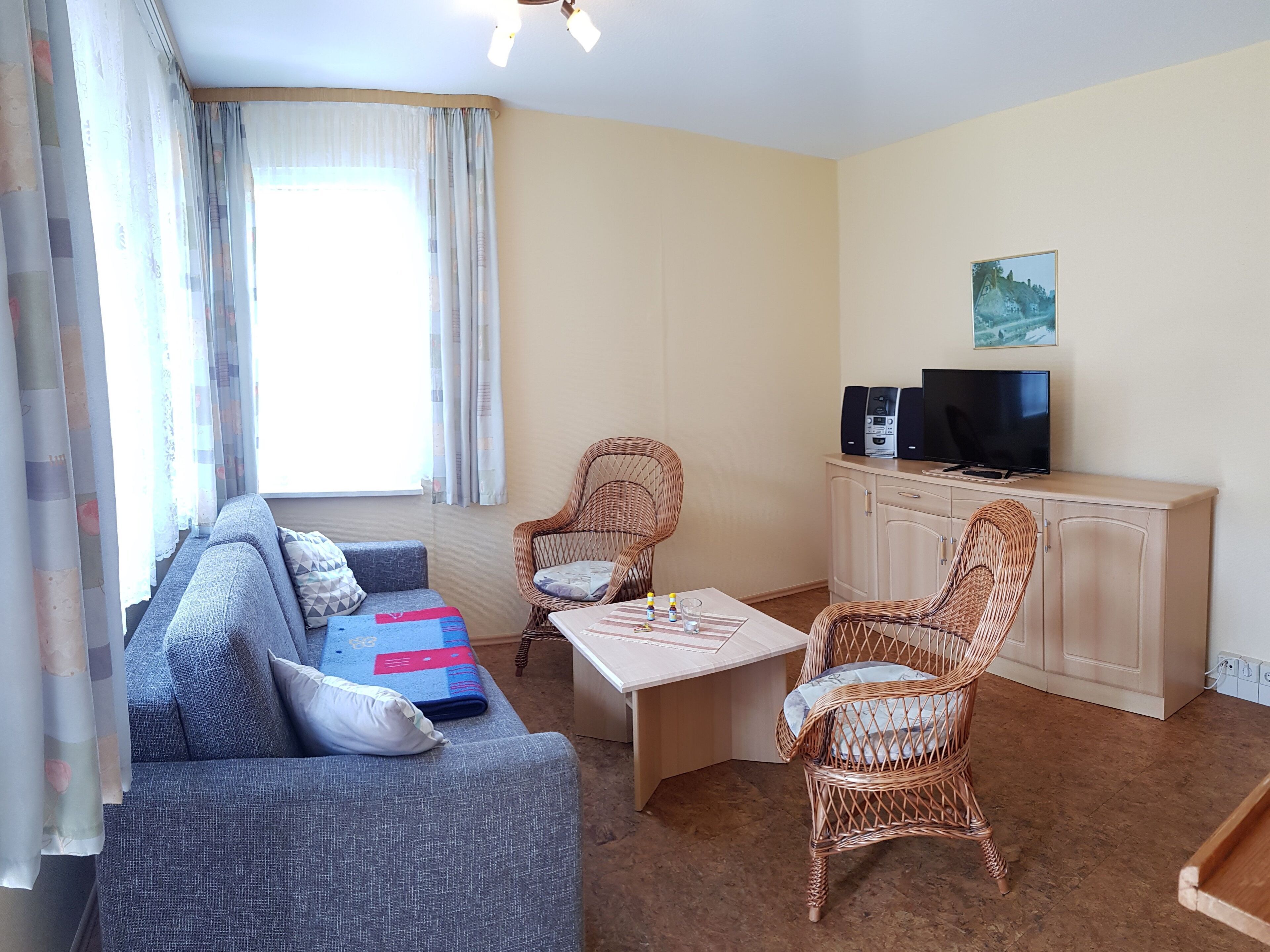 A living room with a sofa, two wicker chairs, a coffee table, a TV on a cabinet, and a window with curtains.