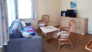 Ein Wohnzimmer mit einer Couch, zwei Korbstühlen, einem Couchtisch, einem Fernseher auf einem Schrank und einem Fenster mit Vorhängen.