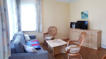 A living room with a sofa, two wicker chairs, a coffee table, a TV on a cabinet, and a window with curtains.