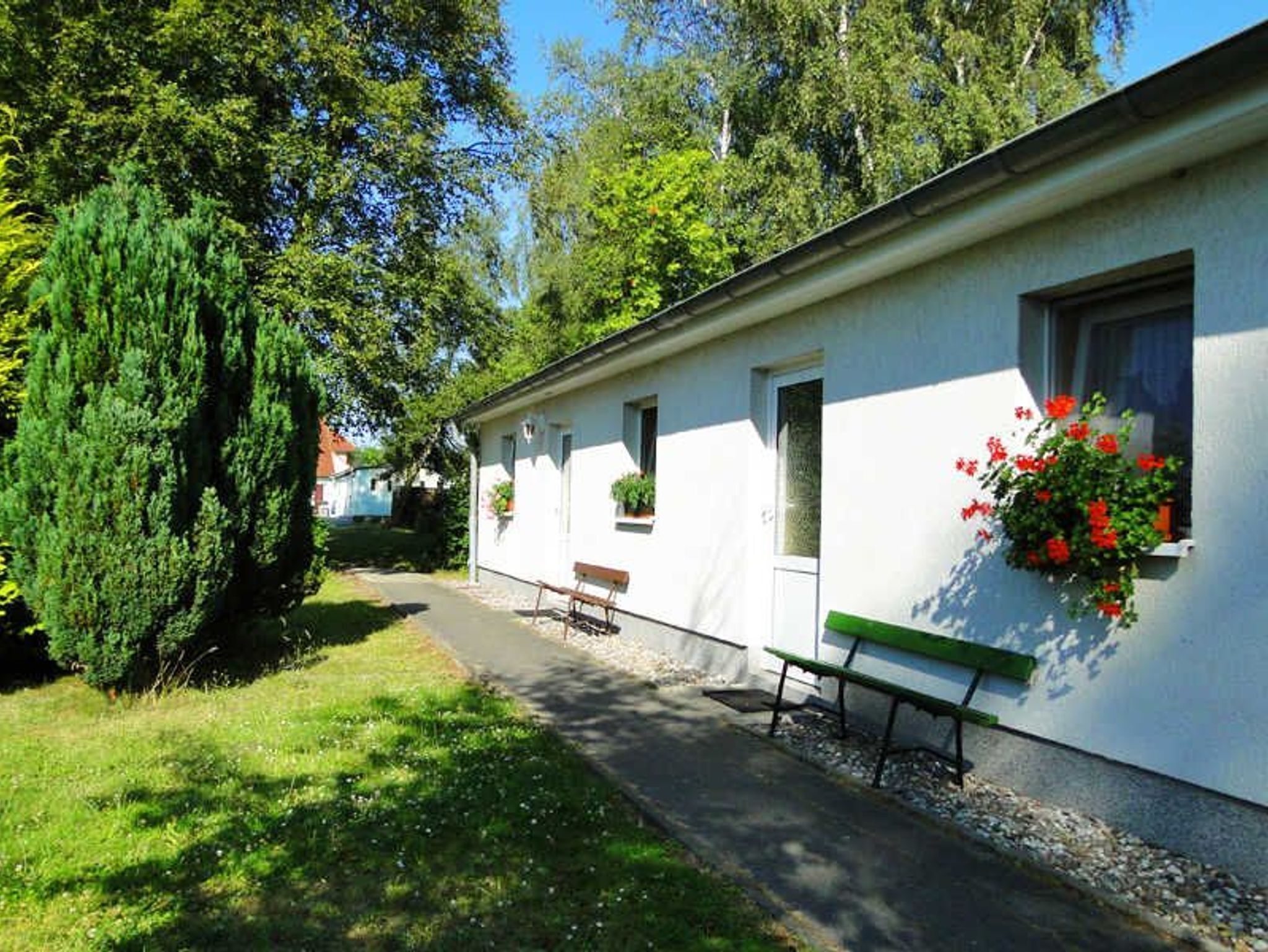 A white building with a bench and a small table outside, surrounded by greenery and trees.