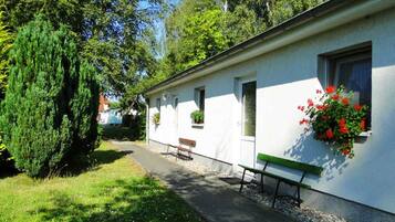 A white building with a bench and a small table outside, surrounded by greenery and trees.