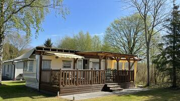 A mobile home with a wooden deck and a covered porch, surrounded by trees.