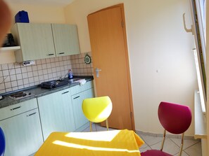 A kitchen with light blue cabinets, a sink, and a microwave. There are two chairs, one yellow and one pink, and a wooden door.