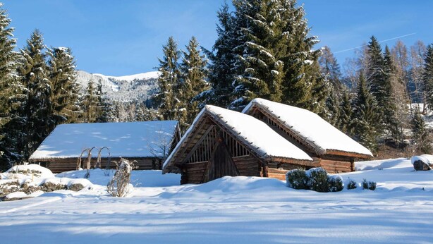 Uma cabana de madeira com telhado coberto de neve, rodeada por árvores e montanhas também cobertas de neve ao fundo.
