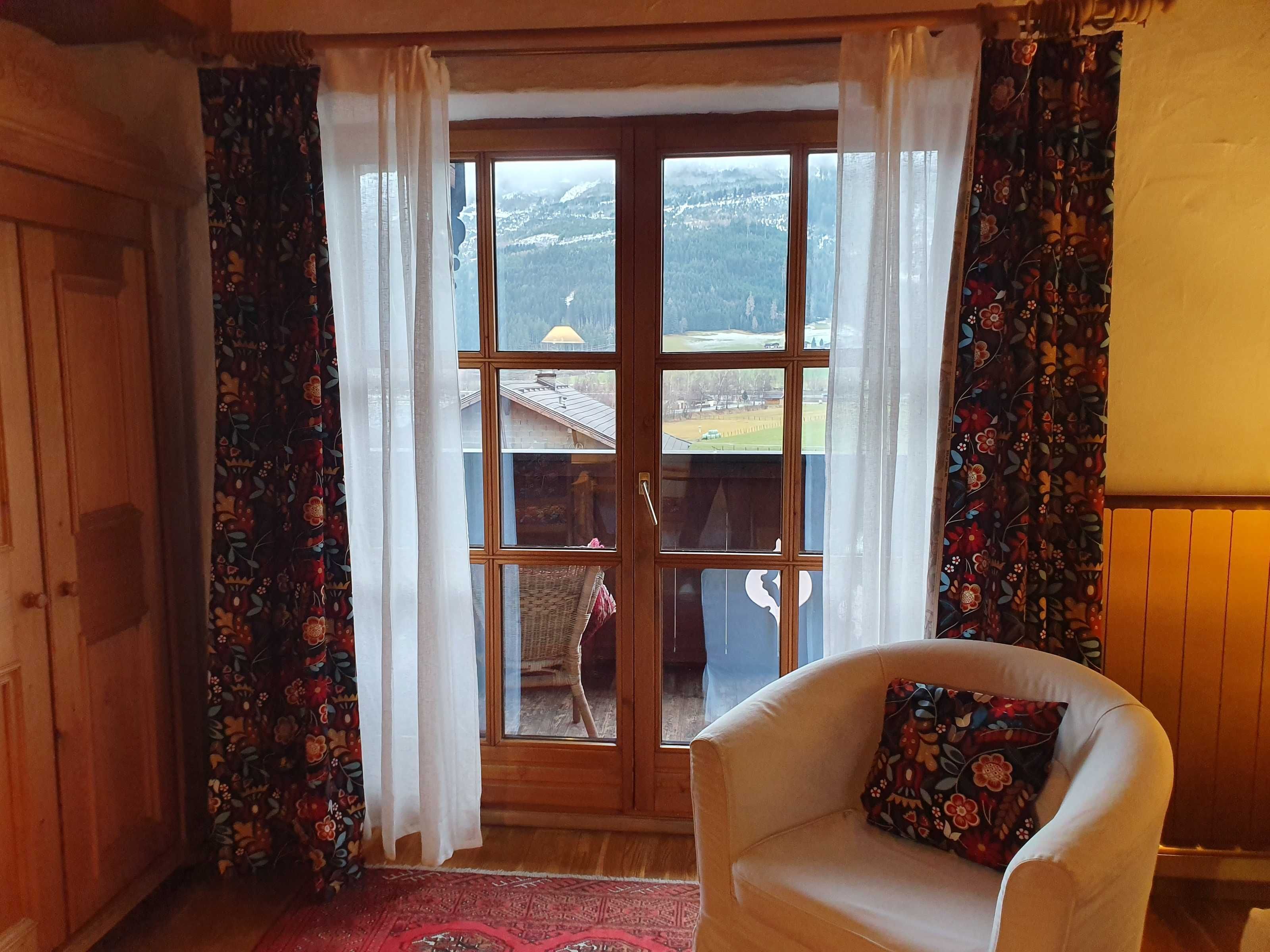 A room with a large window, a wooden door, a white armchair, and a patterned rug.