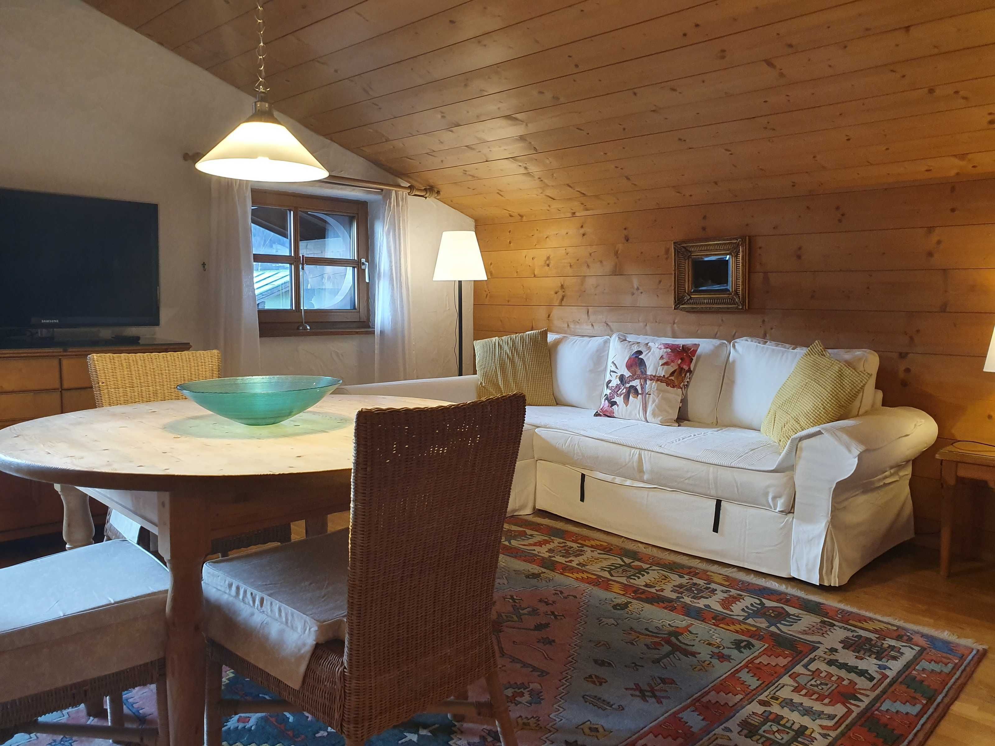 A cozy room with a dining table, wicker chairs, a white sofa, a patterned rug, and wooden walls.