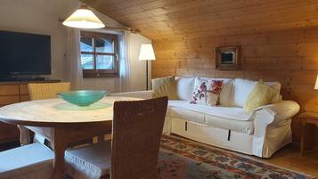 A cozy room with a dining table, wicker chairs, a white sofa, a patterned rug, and wooden walls.