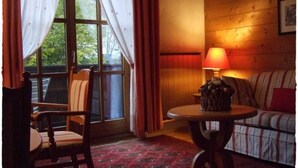 Um quarto acolhedor com piso de madeira, tapete estampado, uma poltrona, um sofá, uma mesa com abajur e uma janela com cortinas.