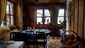 Uma área de refeições com uma mesa posta para dois, uma janela com cortinas e vista para o verde lá fora.