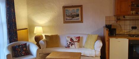 A living room with a sofa, a small table, a lamp, a painting, and a kitchen area with cabinets and a refrigerator.