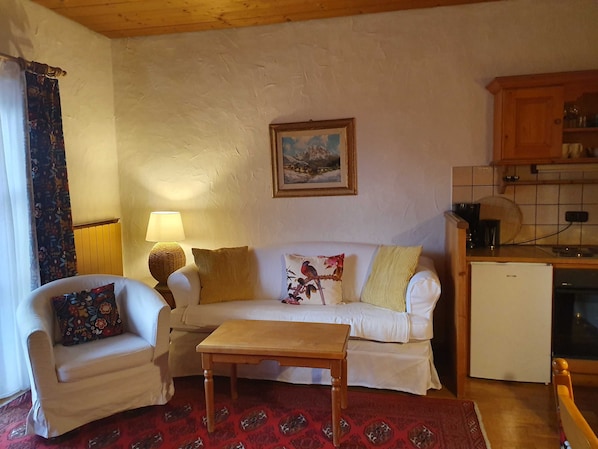 Ein Wohnzimmer mit einer Couch, einem kleinen Tisch, einer Lampe, einem Bild und einer Küchenzeile mit Schränken und einem Kühlschrank.