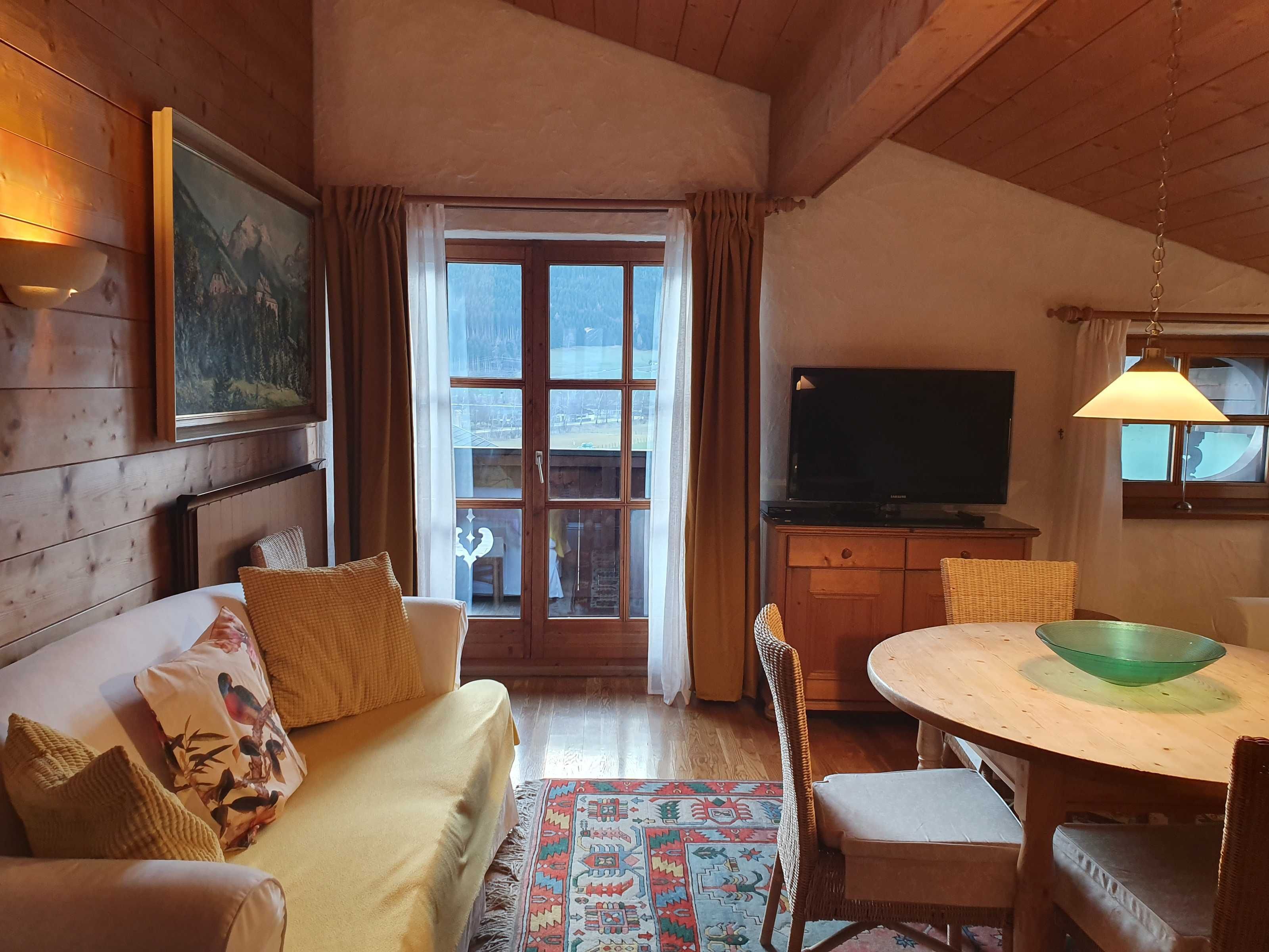 Una habitación acogedora con techo de madera, un sofá con cojines, una mesa de comedor con sillas, un televisor y un amplio ventanal con cortinas.