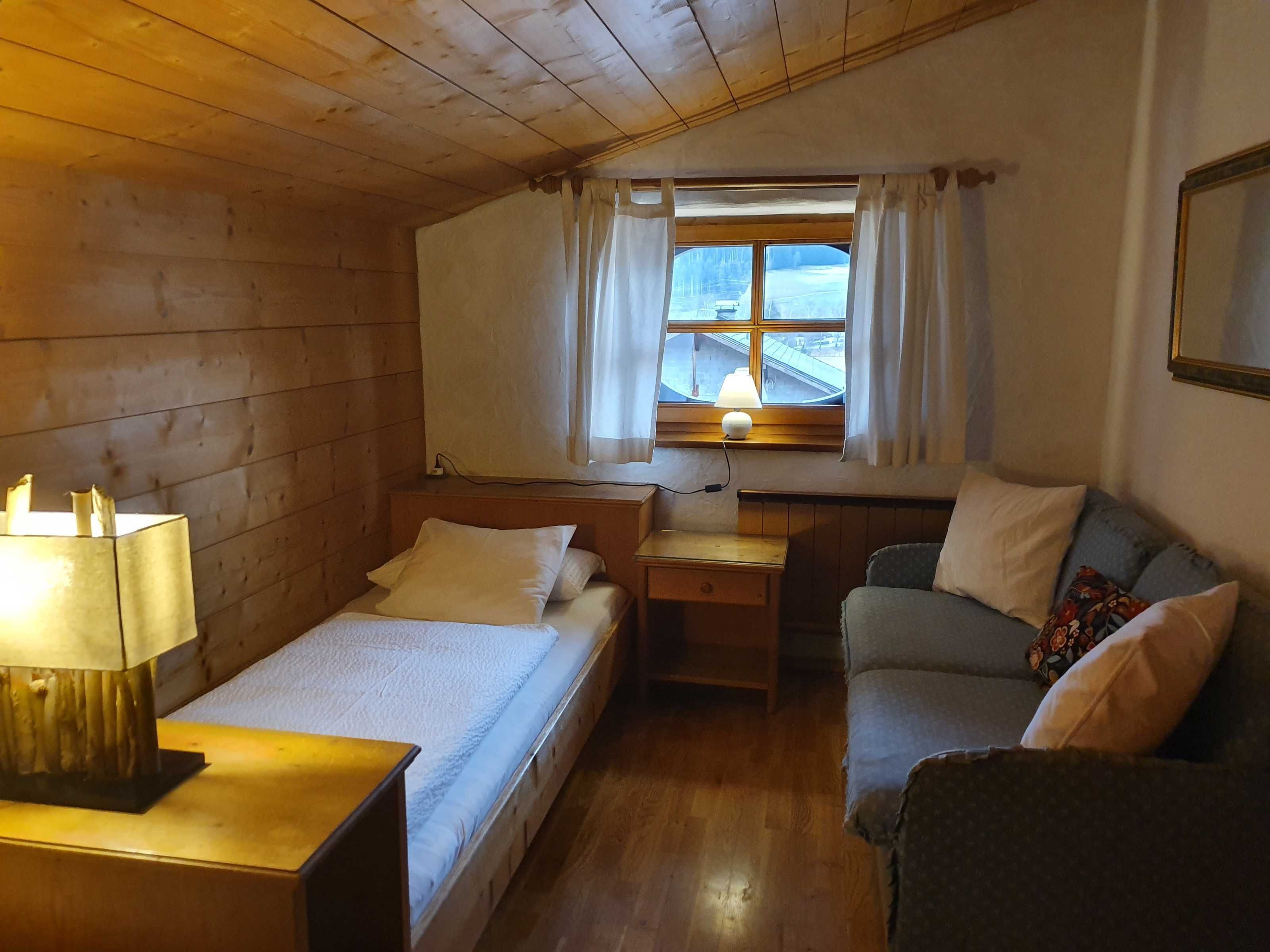 Ein gemütliches Zimmer mit Holzboden, einem Bett mit weißer Bettwäsche, einem kleinen Tisch mit Lampe, einer Couch mit Kissen und einem Fenster mit Vorhängen.