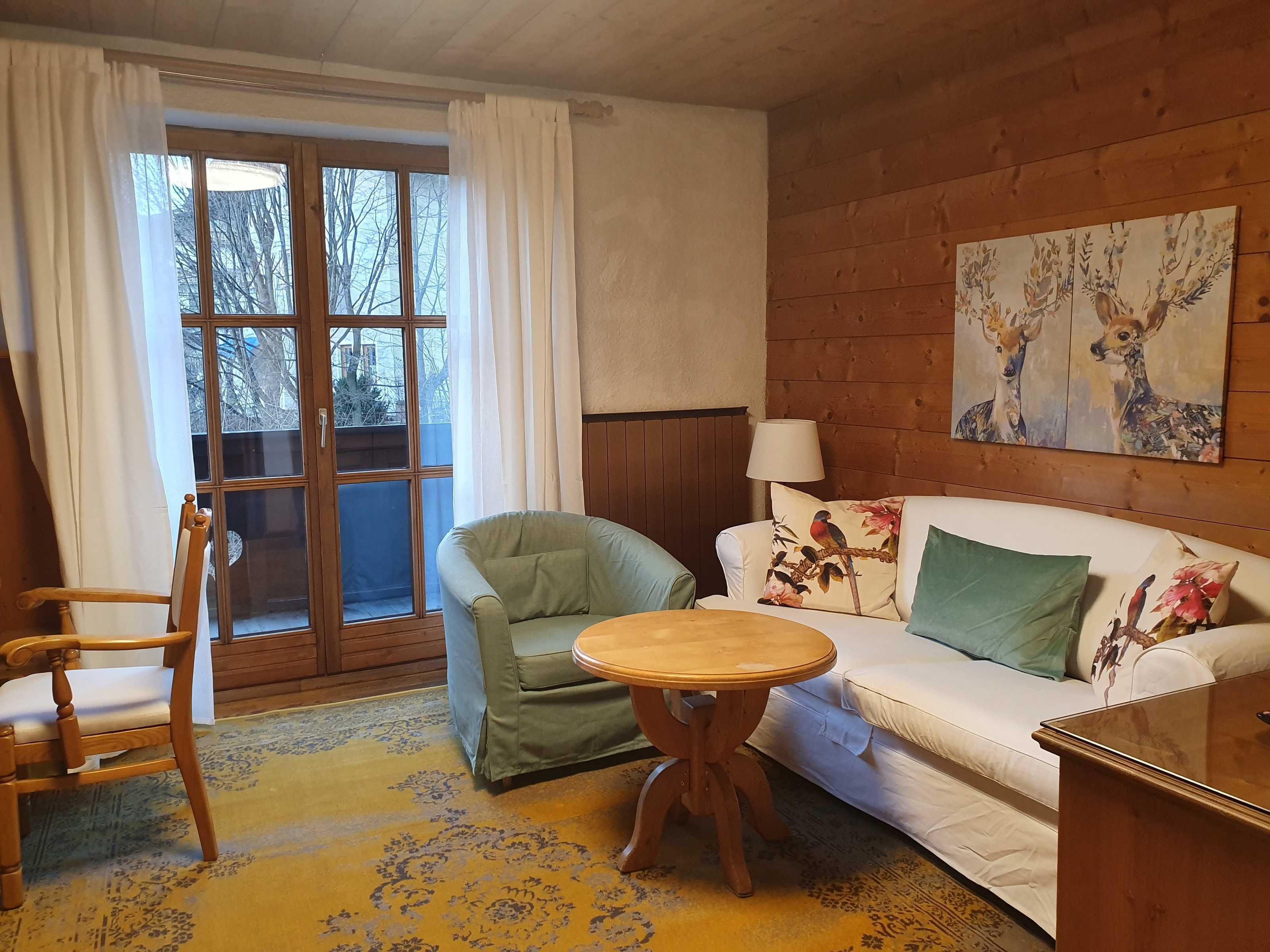Une pièce avec un plancher en bois, un fauteuil vert, un canapé blanc orné de coussins à motifs floraux, une table en bois et un tableau représentant des cerfs accroché au mur.
