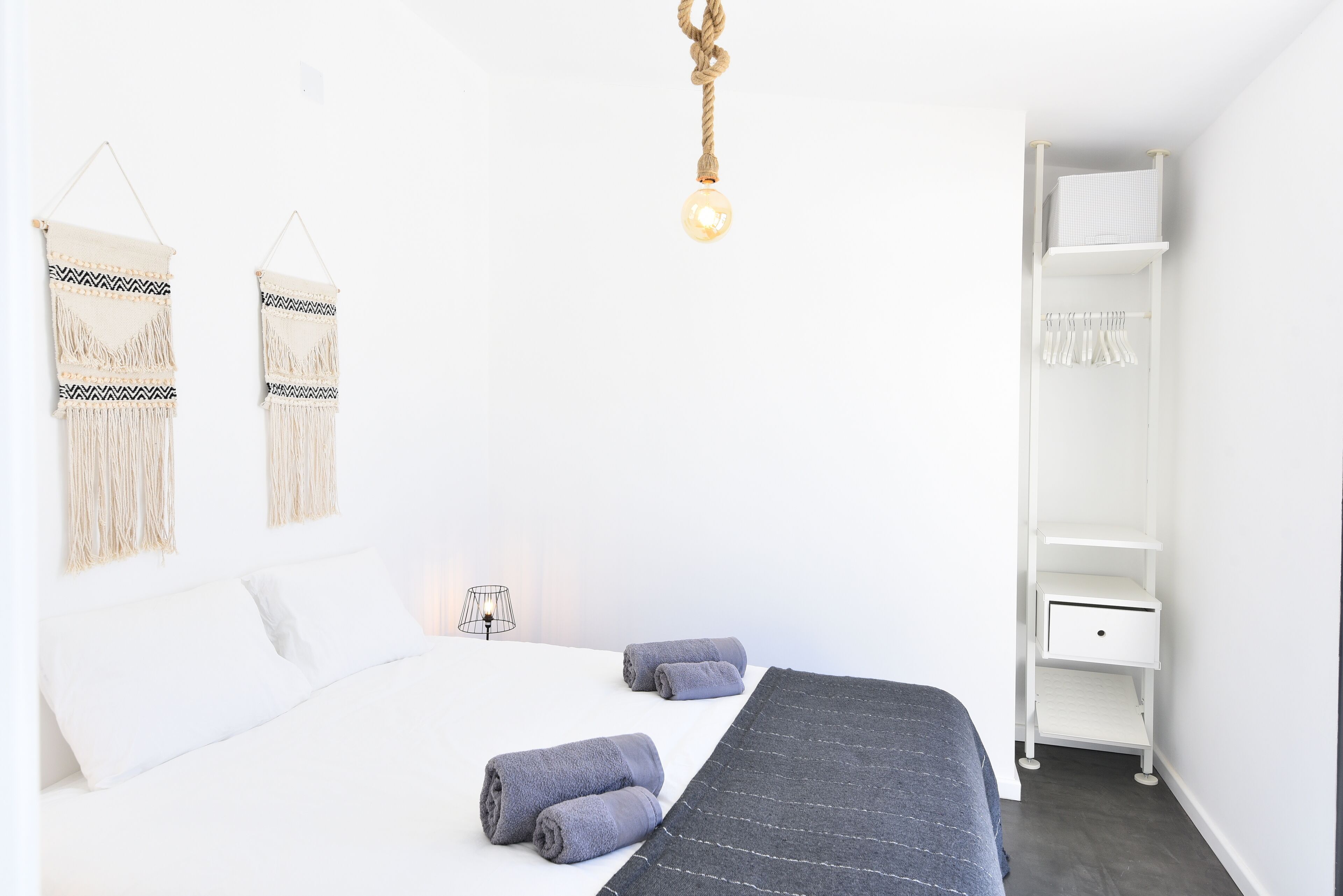 Une chambre à coucher au style minimaliste, avec un lit, deux décorations murales en macramé, un luminaire suspendu et une armoire blanche.