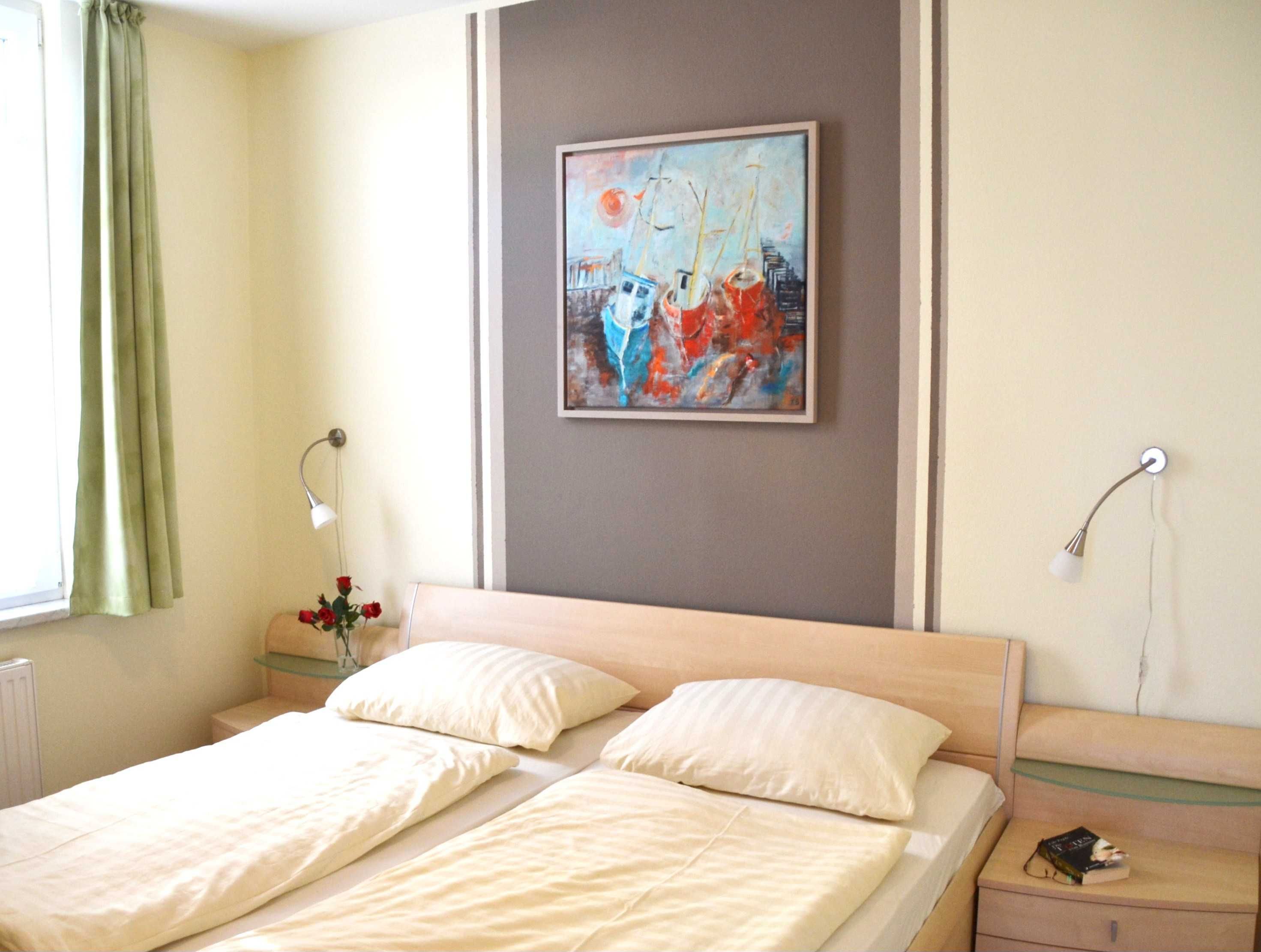 A double bed with two pillows, a bedside table with a lamp, a framed painting on the wall, and a window with curtains.